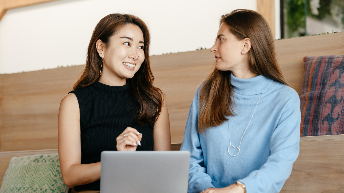 Two women sitting and talking indoors, with one wearing a black sleeveless top and the other wearing a light blue turtleneck sweater. A laptop is in front of them, and cushions and a wooden wall are visible in the background.